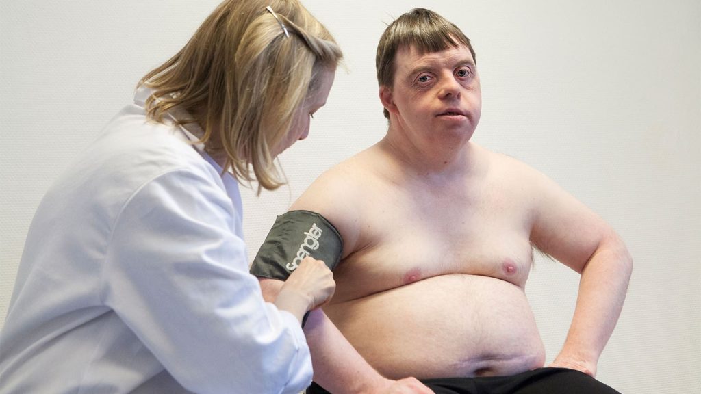 Opinion | Are We Equipped to Care for People With Intellectual, Developmental Disabilities? A female physician fits a blood pressure cuff onto the arm of a man with down syndrome